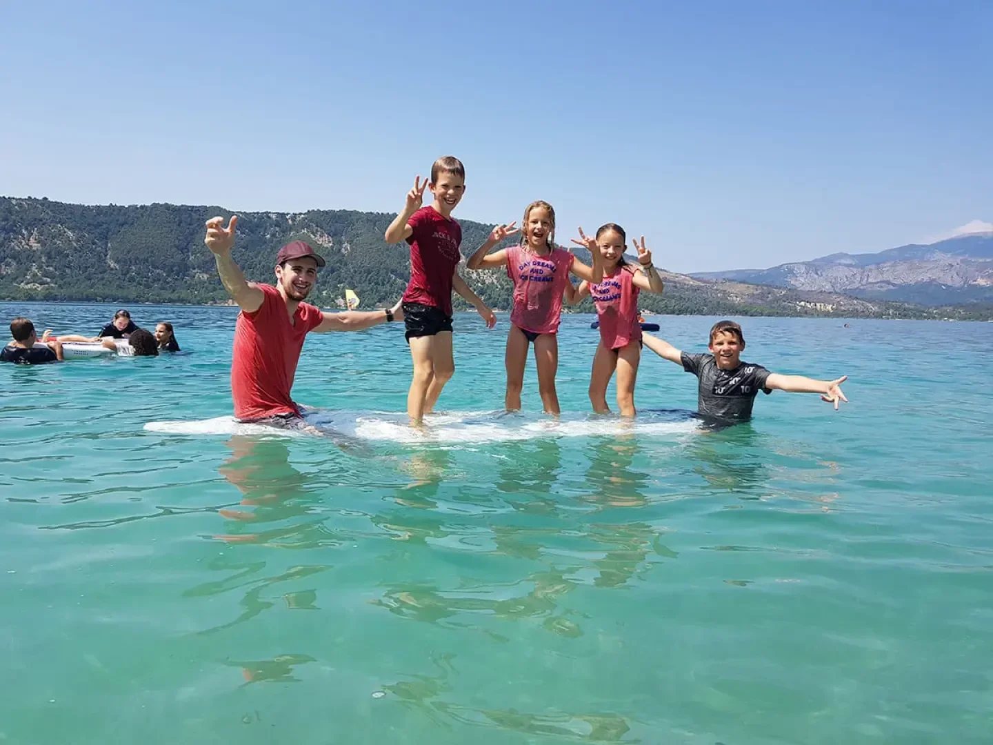 Vier Kinder und ein Erwachsener stehen lächelnd auf einem flachen Felsen in klarem, blauem Wasser und machen Friedenszeichen, während im Hintergrund eine malerische grüne Küste und Berge zu sehen sind. Andere schwimmen in der Nähe im Wasser.