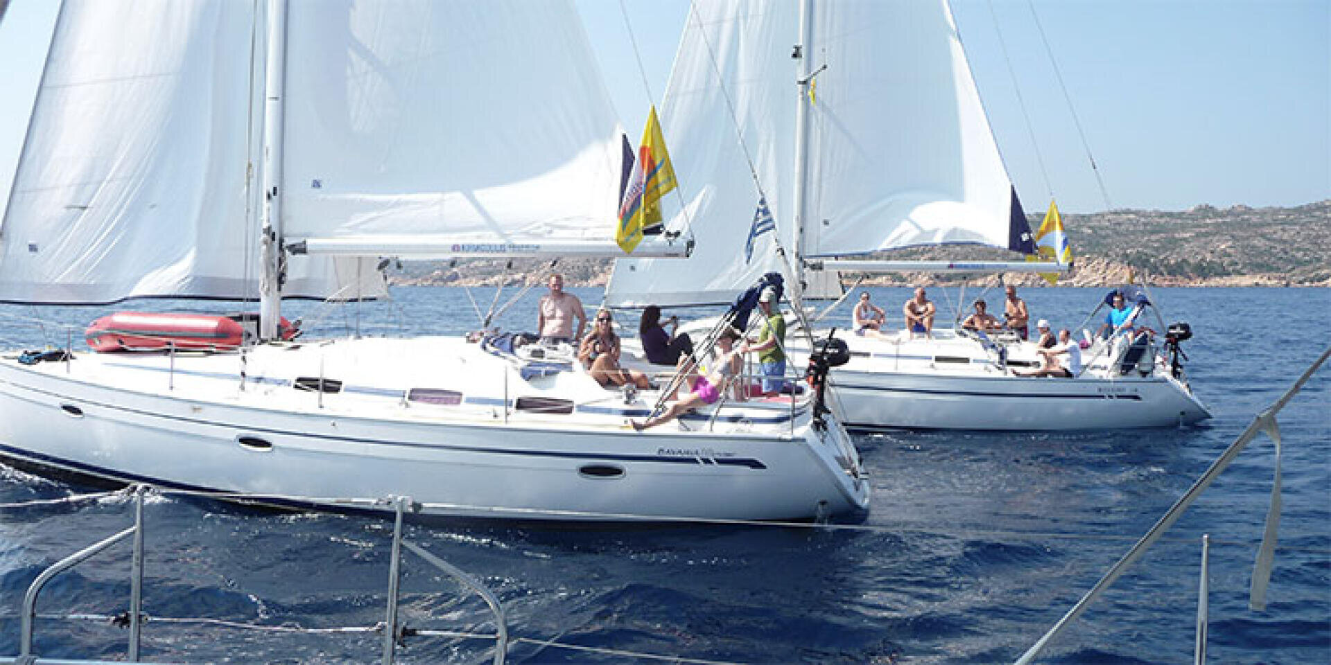 Zwei weiße Segelboote mit Menschen, die sich an Deck sonnen und entspannen, segeln dicht beieinander auf ruhigem, blauem Wasser, mit felsiger Küste und klarem Himmel im Hintergrund.