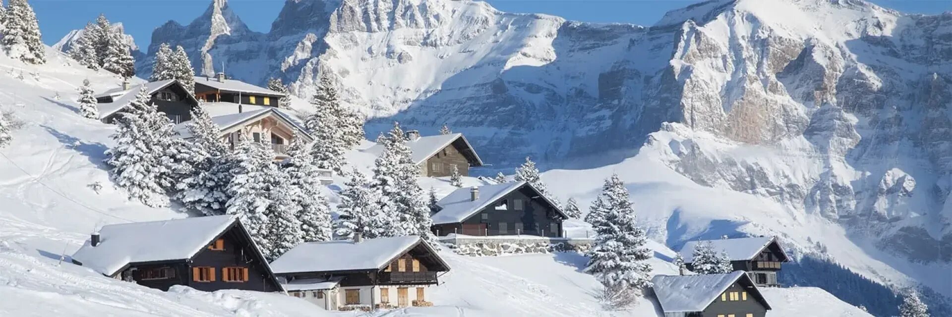 Schneebedeckte Holzhütten inmitten von Kiefern an einem Berghang, mit hohen, schroffen, schneebedeckten Gipfeln und einem klaren blauen Himmel im Hintergrund.