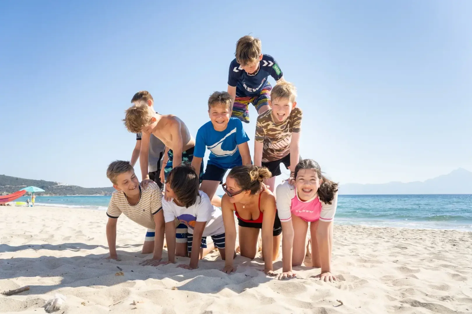 Eine Gruppe von Kindern an einem Sandstrand bildet eine menschliche Pyramide, lächelt und genießt einen sonnigen Tag am Meer. Das Meer und der blaue Himmel sind im Hintergrund zu sehen.