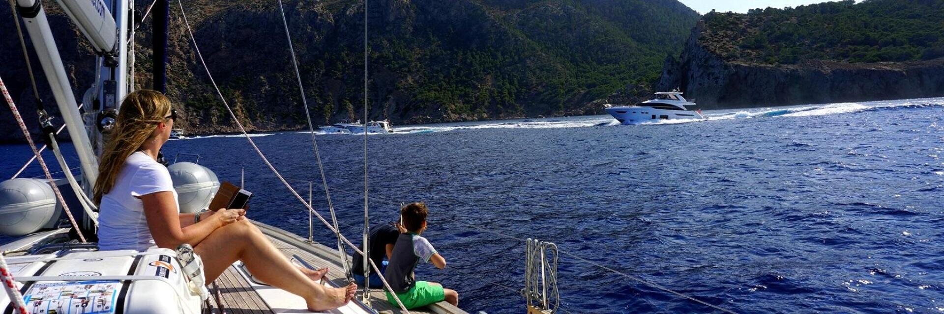 Eine Frau und ein Junge sitzen auf dem Deck eines Segelboots und blicken auf eine große Yacht, die auf dem blauen Meer vorbeifährt, mit felsigen, grünen Hügeln im Hintergrund unter einem klaren Himmel.