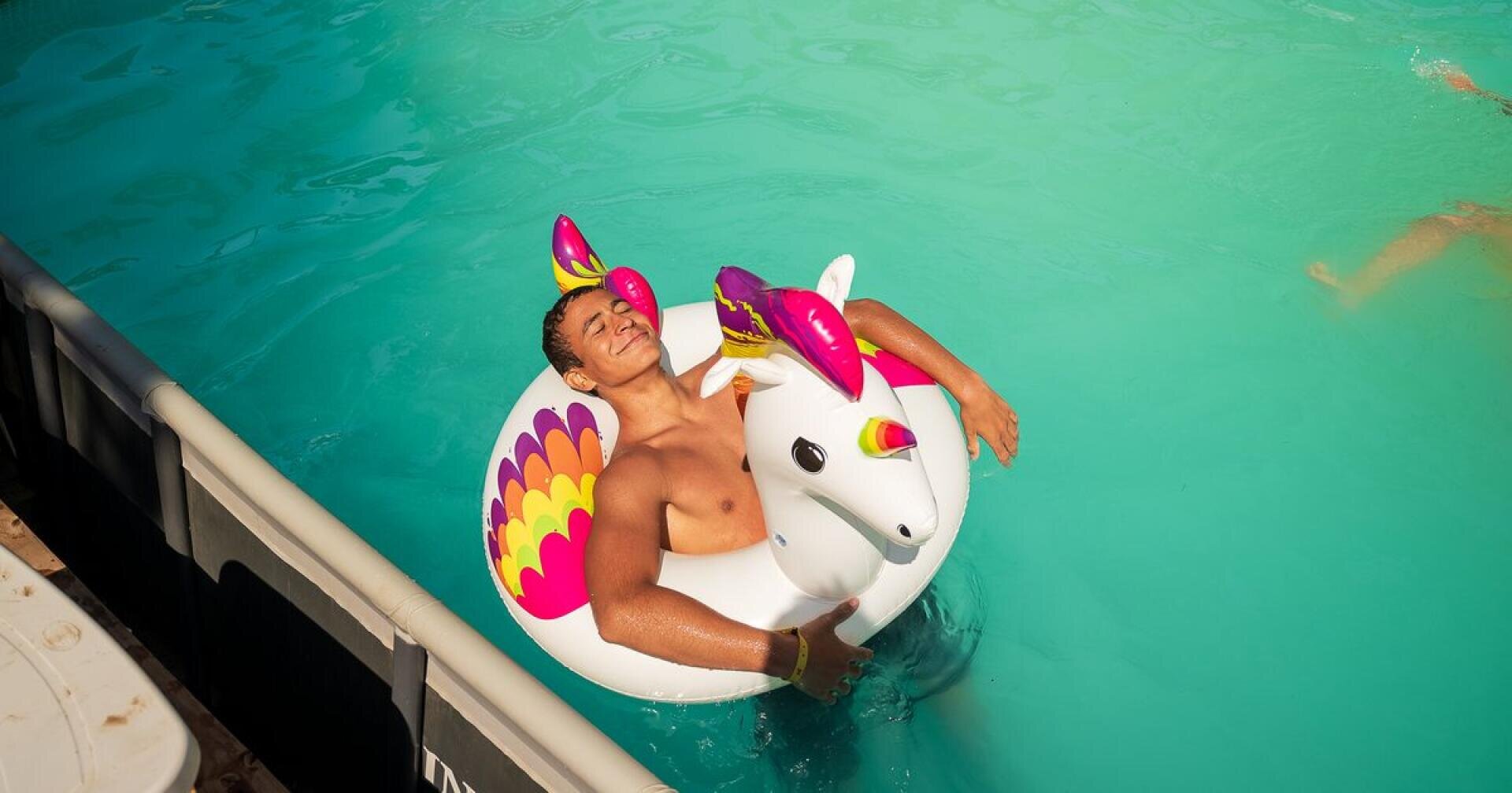 Eine Person entspannt sich in einem Schwimmbecken auf einem bunten aufblasbaren Einhorn-Schwimmring, die Augen geschlossen, während ihr die Sonne ins Gesicht scheint.