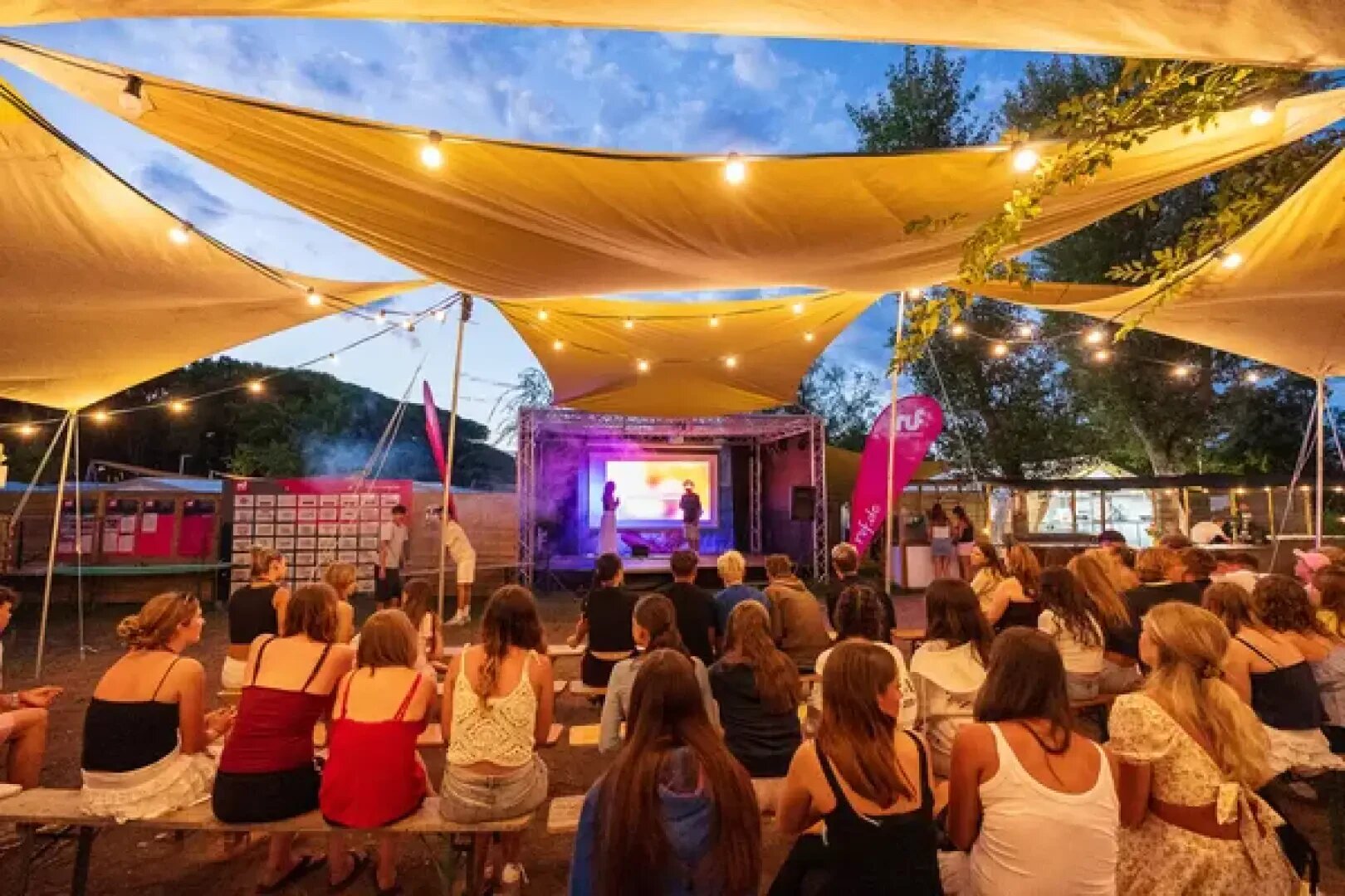 Ein Publikum sitzt auf Bänken unter gelben Baldachinen und verfolgt eine Präsentation auf einer Freilichtbühne in der Abenddämmerung. Über dem Kopf hängen Lichterketten, und im Hintergrund sind Bäume und ein bunter Himmel zu sehen.