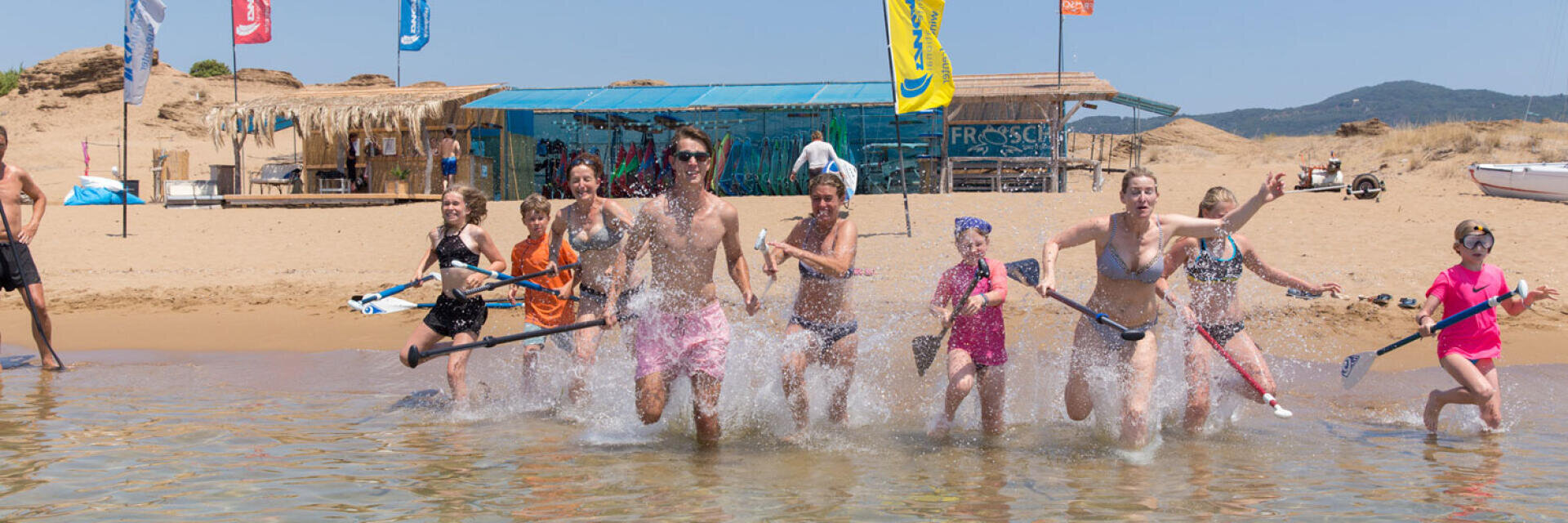 Eine Gruppe von Menschen an einem Strand, die Paddel in der Hand halten und ins flache Wasser laufen, planschen und lächeln, mit einer Strandhütte, Flaggen und Hügeln im Hintergrund unter einem klaren Himmel.