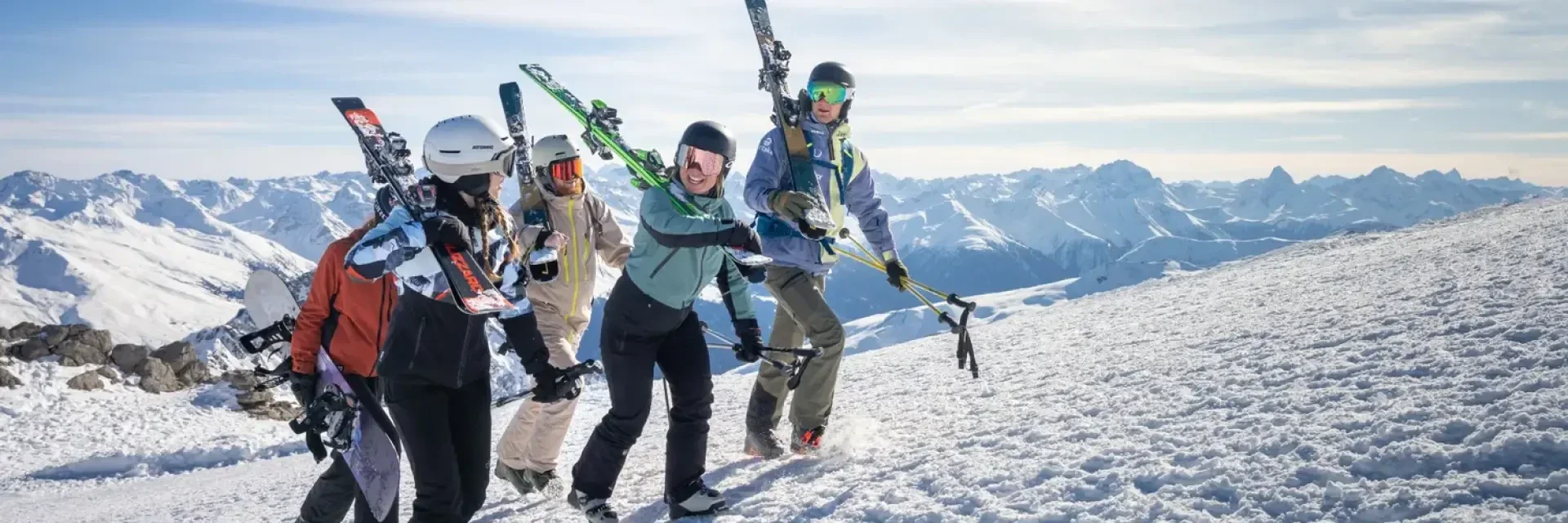 Eine Gruppe von fünf Skifahrern in Winterausrüstung, die ihre Skier tragen, geht einen verschneiten Hang hinauf, mit schneebedeckten Bergen im Hintergrund und unter einem klaren Himmel.