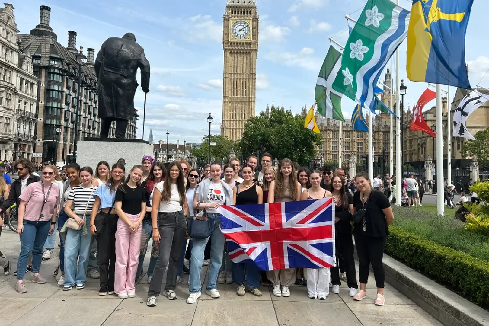 Eine Gruppe von Menschen posiert mit einer großen Union-Jack-Flagge auf einem Stadtplatz. Im Hintergrund sind an einem teilweise bewölkten Tag Flaggen, eine Statue und der Big Ben zu sehen.