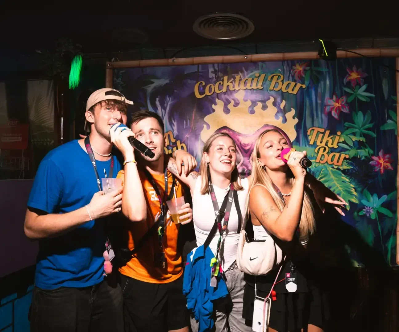 Vier junge Erwachsene singen gemeinsam Karaoke in einer farbenfrohen Bar mit einem Schild "Cocktail Bar" und "Tiki Bar" im Hintergrund. Sie sehen fröhlich und energiegeladen aus, halten Mikrofone und Getränke in der Hand und stehen unter buntem Licht.