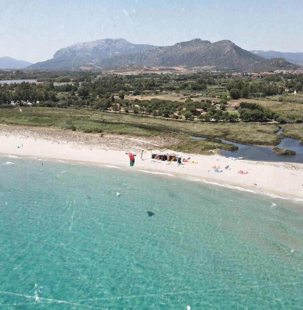 Luftaufnahme eines Sandstrandes mit türkisfarbenem Wasser, ein paar bunten Sonnenschirmen und Menschen, die sich in der Nähe des Ufers entspannen. Im Hintergrund sind grüne Felder, Bäume und Berge unter einem klaren Himmel zu sehen.