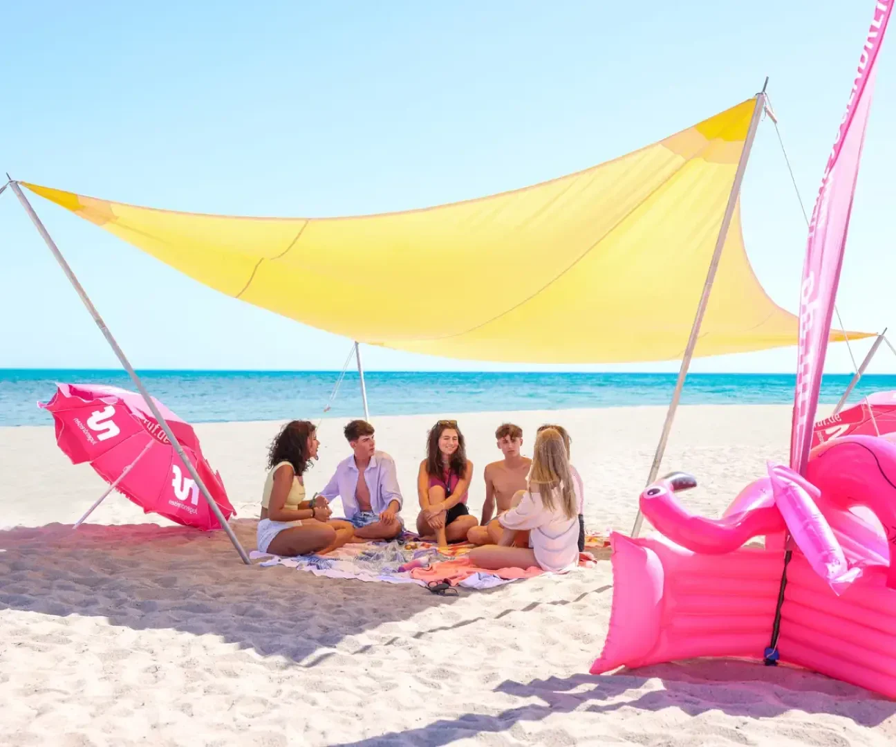 Fünf Personen sitzen auf Handtüchern unter einem großen gelben Vordach an einem Sandstrand, unterhalten sich und genießen das sonnige Wetter. Rosa aufblasbare Poolspielzeuge und Flaggen sind in der Nähe, das Meer ist im Hintergrund zu sehen.