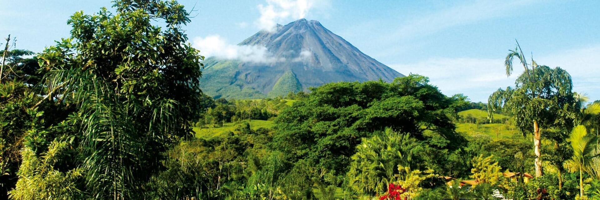 Eine üppig grüne tropische Landschaft mit dichten Bäumen und Pflanzen im Vordergrund und einem großen vulkanischen Berg, der teilweise mit Wolken bedeckt ist und sich im Hintergrund unter einem strahlend blauen Himmel erhebt.