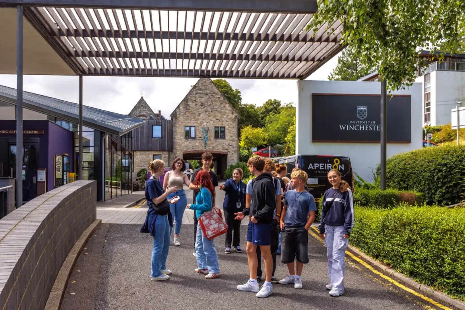 Eine Gruppe junger Leute steht an einem sonnigen Tag vor den Gebäuden der Universität von Winchester und unterhält sich, umgeben von Bäumen und Grünpflanzen und einem großen Schild mit dem Namen der Universität im Hintergrund.