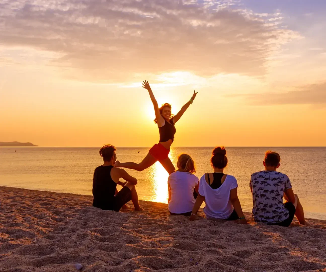 Eine Person springt freudig mit erhobenen Armen auf einem Sandstrand bei Sonnenuntergang, während vier andere auf dem Sand sitzen und auf das ruhige Meer und den bunten Himmel blicken.