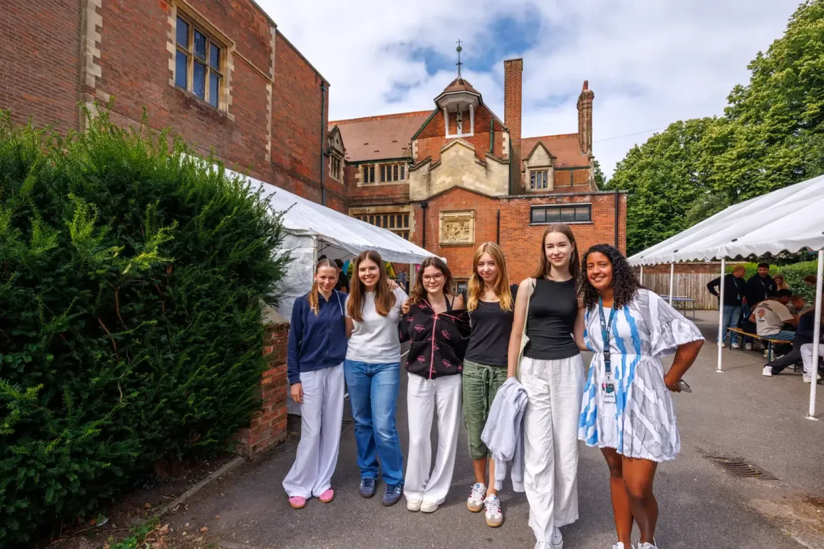 An einem sonnigen Tag mit teilweise bewölktem Himmel stehen sechs junge Frauen lächelnd vor einem roten Backsteingebäude mit weißen Zelten daneben.