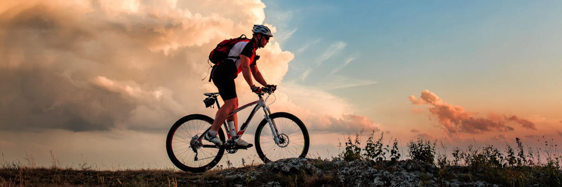 Eine Person mit Helm und Rucksack fährt mit dem Mountainbike auf einem felsigen Weg bei Sonnenuntergang, mit dramatischen Wolken und einem bunten Himmel im Hintergrund.