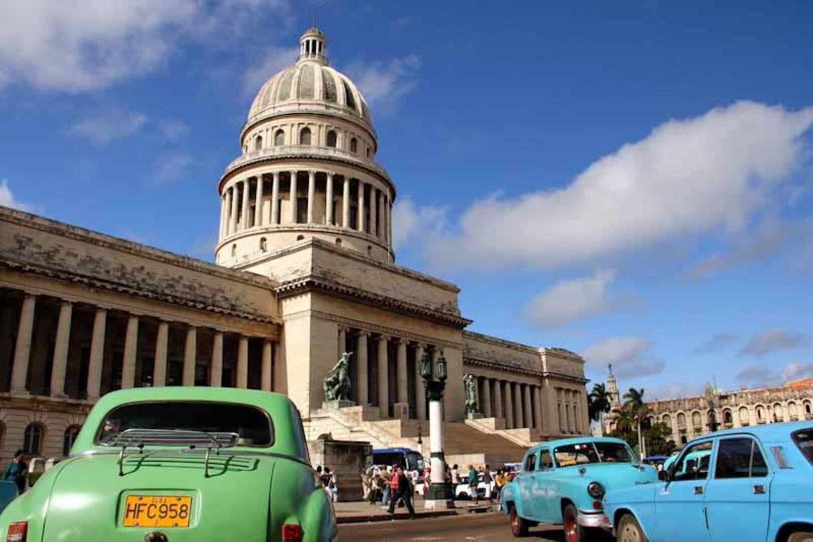 Ein Blick auf El Capitolio, ein großes Kuppelgebäude in Havanna, Kuba, mit klassischen Oldtimern in Grün und Blau im Vordergrund unter einem blauen Himmel mit vereinzelten Wolken.