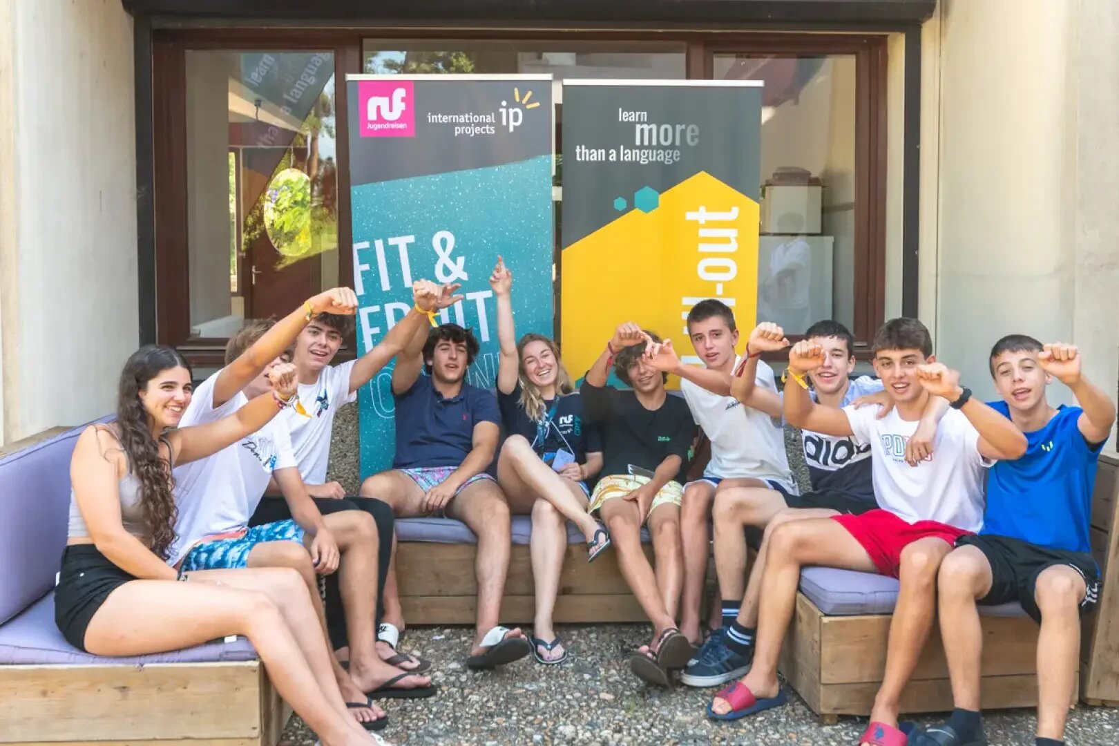Eine Gruppe von acht lächelnden Teenagern sitzt auf Sofas im Freien, hebt die Arme und macht Daumen-hoch-Gesten. Hinter ihnen hängen bunte Banner mit den Aufschriften "FIT & FUN" und "Learn more than a language".