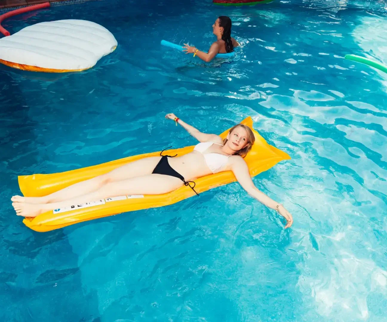 Eine Frau im Bikini liegt auf einem gelben aufblasbaren Schwimmkörper und entspannt sich in einem blauen Schwimmbecken. Im Hintergrund schwimmt eine andere Person, die eine Poolnudel hält.