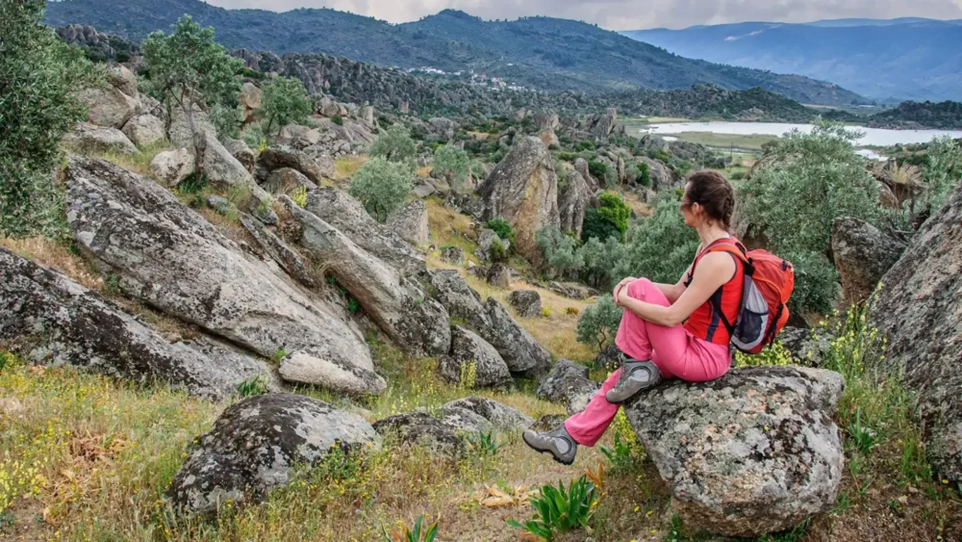 Eine Person, die einen roten Rucksack und eine rosa Hose trägt, sitzt auf einem Felsen und blickt auf eine felsige, grüne Landschaft mit verstreuten Bäumen, Hügeln und einem entfernten Gewässer unter einem bewölkten Himmel.