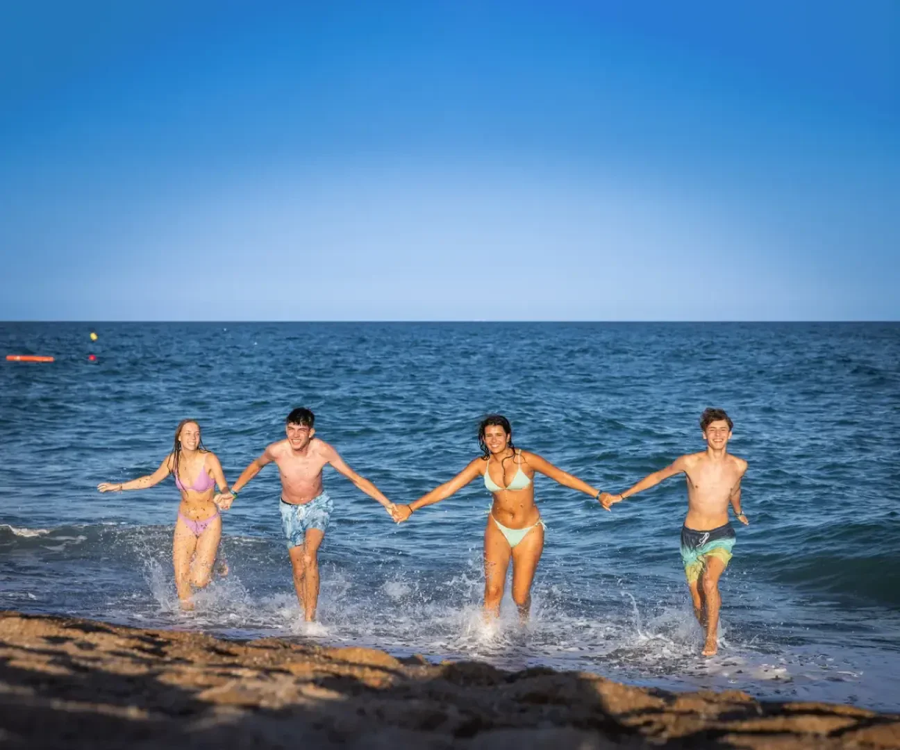 Vier junge Erwachsene in Badeanzügen halten sich an den Händen und laufen aus dem Meer auf einen Sandstrand, wo sie mit dem klaren blauen Himmel und dem Meer im Hintergrund planschen.