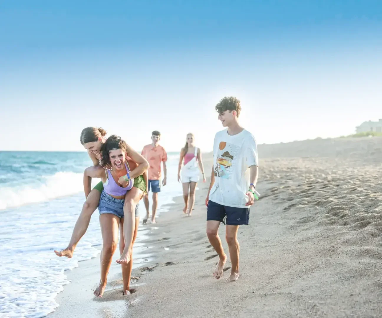 Eine Gruppe junger Leute spaziert und spielt an einem Sandstrand in der Nähe des Ozeans. Eine Person nimmt eine andere huckepack, während die anderen lächelnd spazieren gehen und den sonnigen Tag genießen. Der Himmel ist klar und blau.