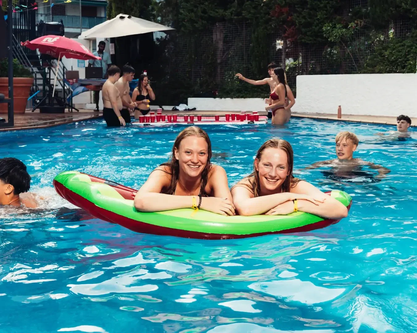Zwei junge Frauen liegen lächelnd auf einem aufblasbaren Schwimmkörper in einem Schwimmbad. Im Hintergrund schwimmen andere Menschen und spielen Spiele an einem sonnigen Tag.