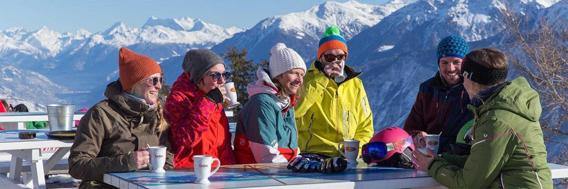 Eine Gruppe von sechs Personen in Winterkleidung sitzt an einem Tisch im Freien mit heißen Getränken, lacht und unterhält sich vor der Kulisse verschneiter Berge und strahlend blauem Himmel. Auf dem Tisch liegen Skiausrüstungen, darunter Helme und Handschuhe.