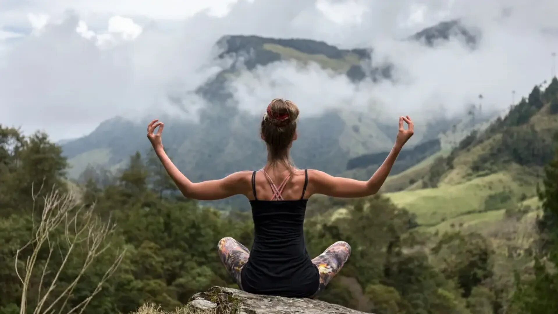 Eine Frau sitzt im Schneidersitz auf einem Felsen und meditiert mit ihren Händen in einer Mudra-Position. Sie blickt auf eine neblige Berglandschaft mit grünen Hügeln und Bäumen.