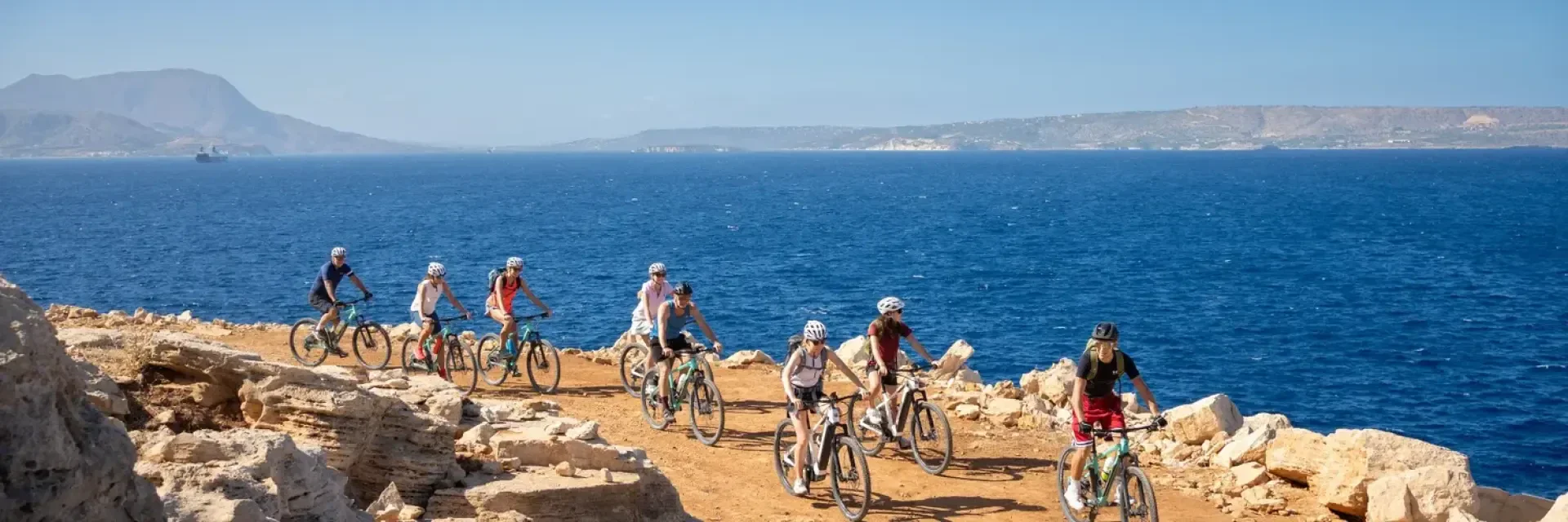 Eine Gruppe von Menschen mit Helmen fährt mit dem Fahrrad entlang einer felsigen Küstenklippe mit klarem blauen Meer und fernen Bergen unter einem hellen, wolkenlosen Himmel.