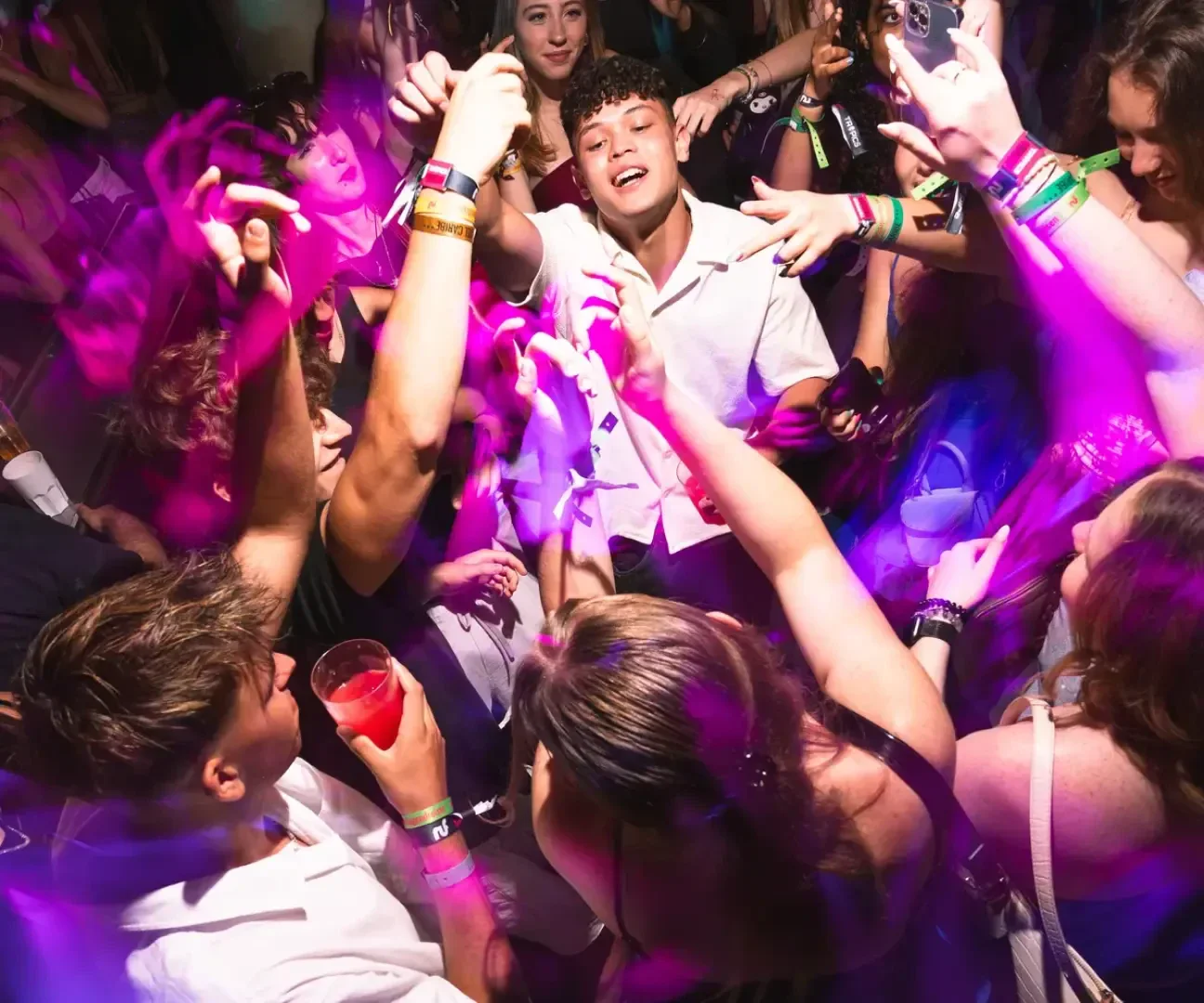 Eine Gruppe junger Leute tanzt eng beieinander auf einer Party, lächelt und hebt die Hände. Lila und rosa Lichter schaffen eine lebhafte, energiegeladene Atmosphäre. Einige halten Getränke in der Hand und tragen Armbänder, um die festliche Nacht zu genießen.