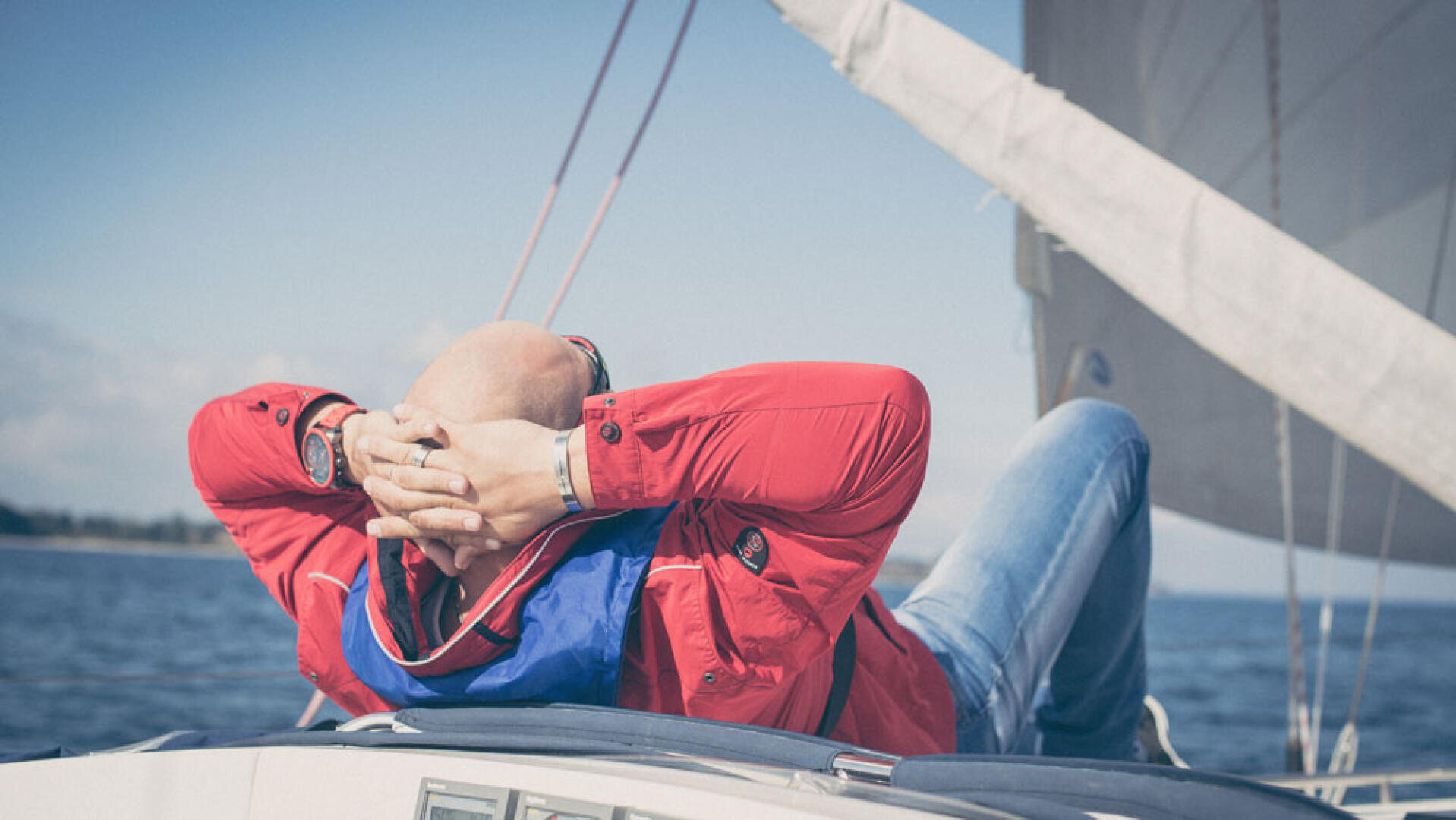 Eine Person in roter Jacke und blauer Jeans entspannt sich mit den Händen hinter dem Kopf auf einem Segelboot und genießt einen sonnigen Tag auf dem Wasser, wobei das Segel im Hintergrund sichtbar ist.