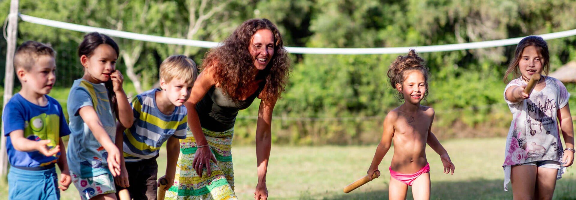 Eine Frau und fünf kleine Kinder stehen im Freien auf einer Wiese und bereiten sich auf den Start eines Rennens vor. Die Kinder lächeln und halten Schlagstöcke, im Hintergrund sind Bäume und ein Volleyballnetz zu sehen, ein sonniger Tag.