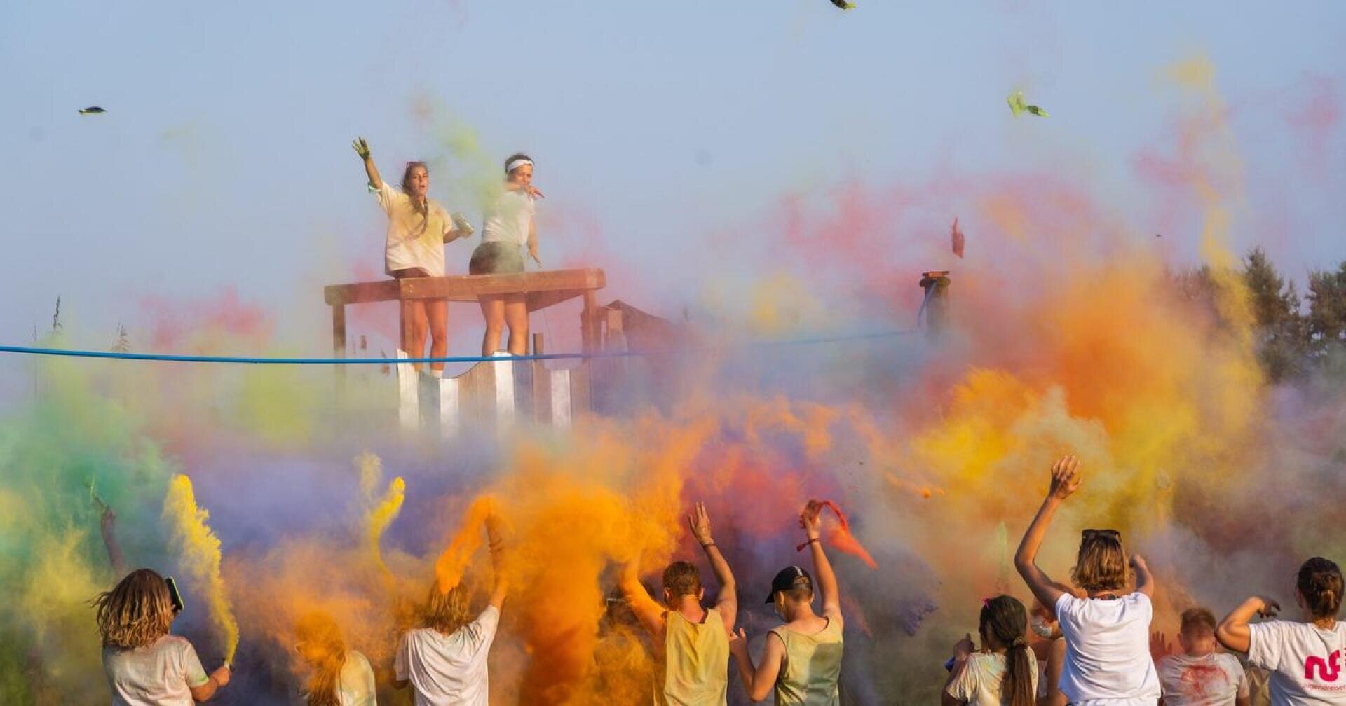 Eine Menschenmenge wirft buntes Pulver in die Luft, während zwei Personen auf einer Plattform darüber stehen und bei strahlendem Himmel auf einem Open-Air-Festival feiern.