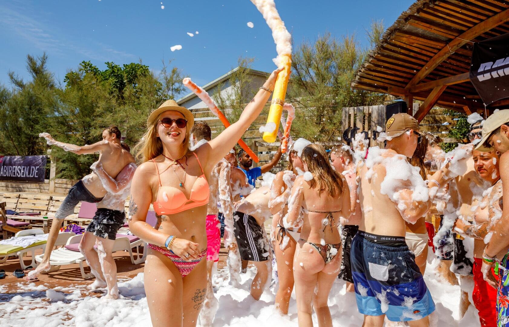 Eine Gruppe von Menschen in Badeanzügen genießt eine Schaumparty im Freien unter sonnigem Himmel. Eine lächelnde Frau mit Sonnenbrille und Hut versprüht Schaum, während andere in den Blasen um sie herum tanzen und spielen.