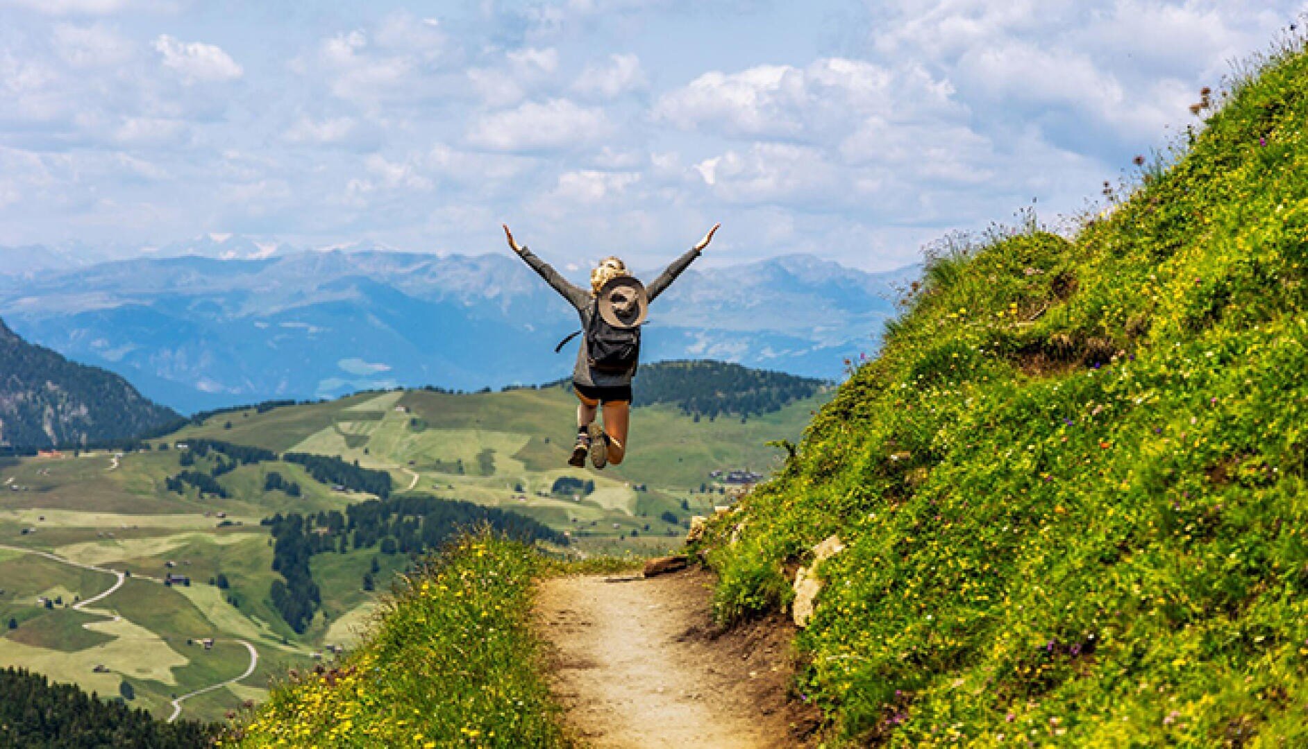Eine Person mit einem Rucksack springt freudig auf einem Bergpfad, umgeben von grünen Hügeln und fernen Bergen unter einem teilweise bewölkten Himmel.