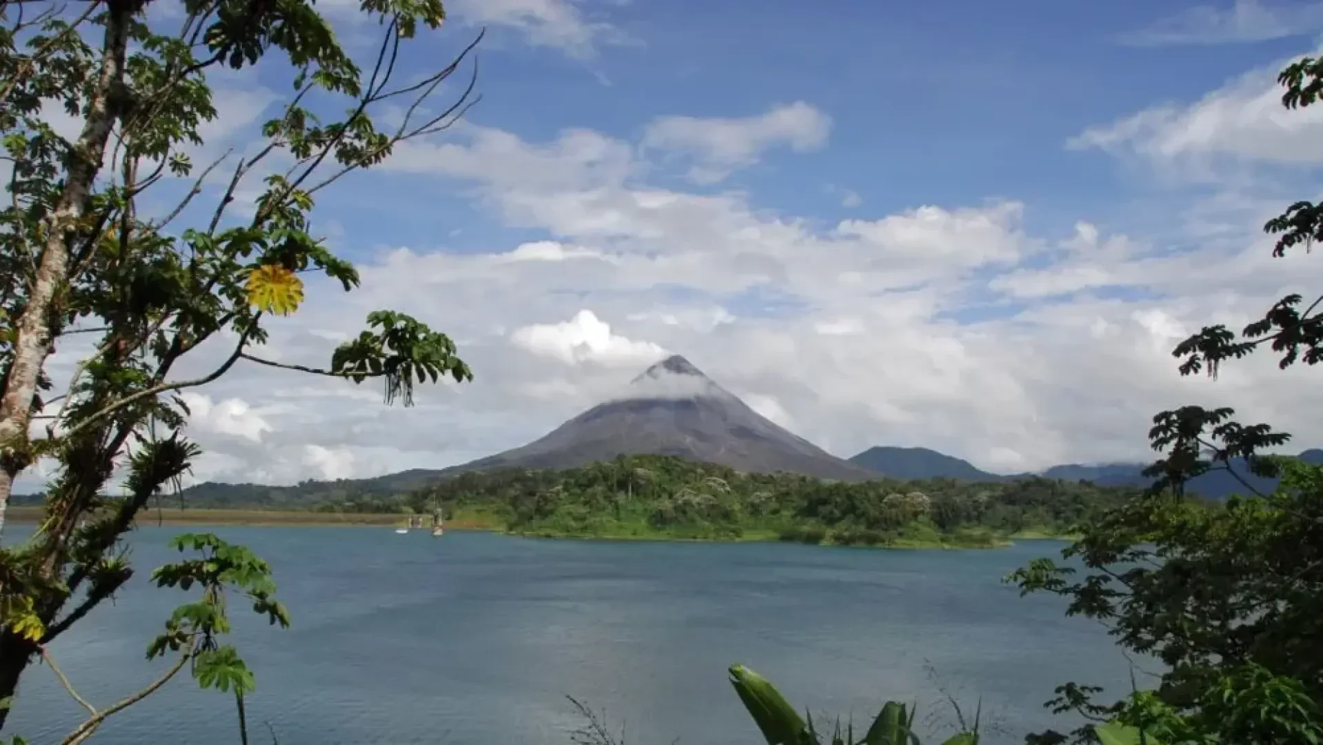 Fernsicht auf einen kegelförmigen Vulkan, umgeben von üppigem Grün, mit einem See im Vordergrund und einem teilweise bewölkten blauen Himmel über dem Kopf, eingerahmt von Baumzweigen.