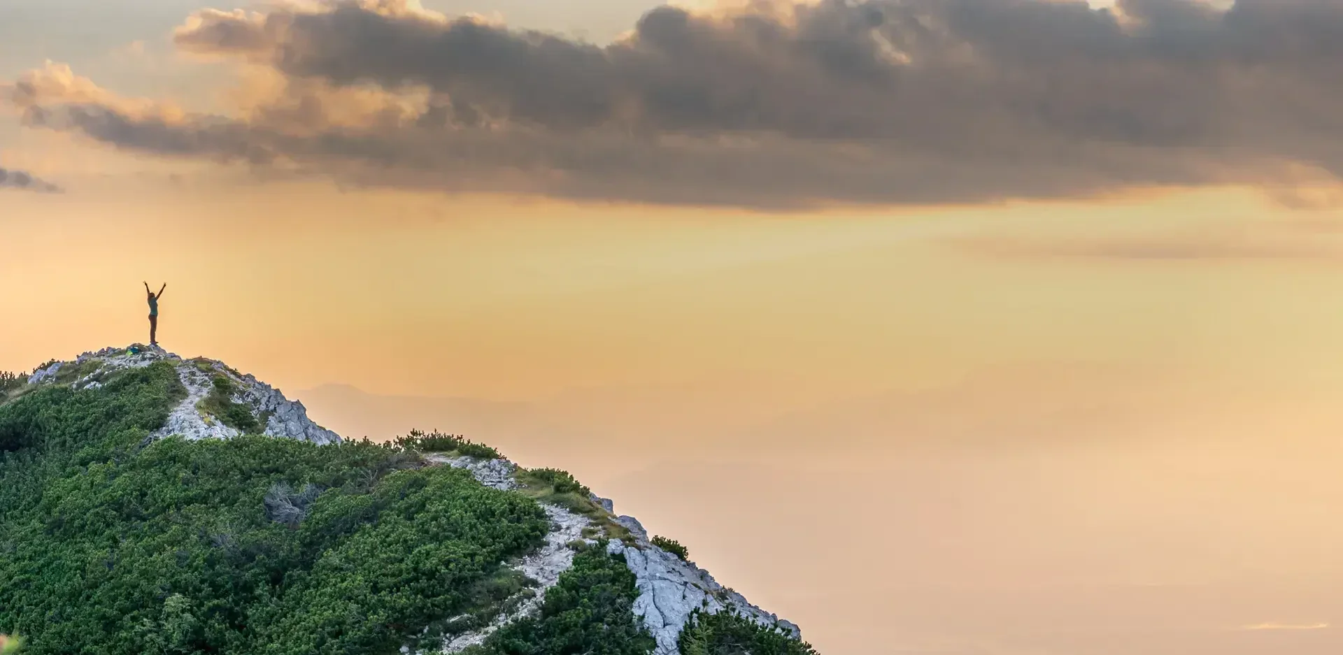 Eine Person steht mit siegreich erhobenen Armen auf einem felsigen, grünen Hügel bei Sonnenaufgang oder Sonnenuntergang, unter einem Himmel mit weichen Wolken und warmem, goldenem Licht.