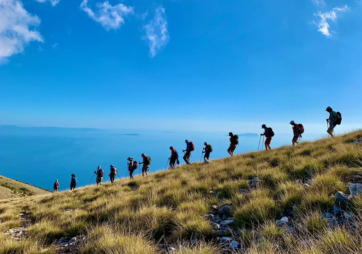 Eine Gruppe von Wanderern mit Rucksäcken wandert auf einem grasbewachsenen Hang unter einem strahlend blauen Himmel bergauf, während im Hintergrund das Meer und der ferne Horizont zu sehen sind.