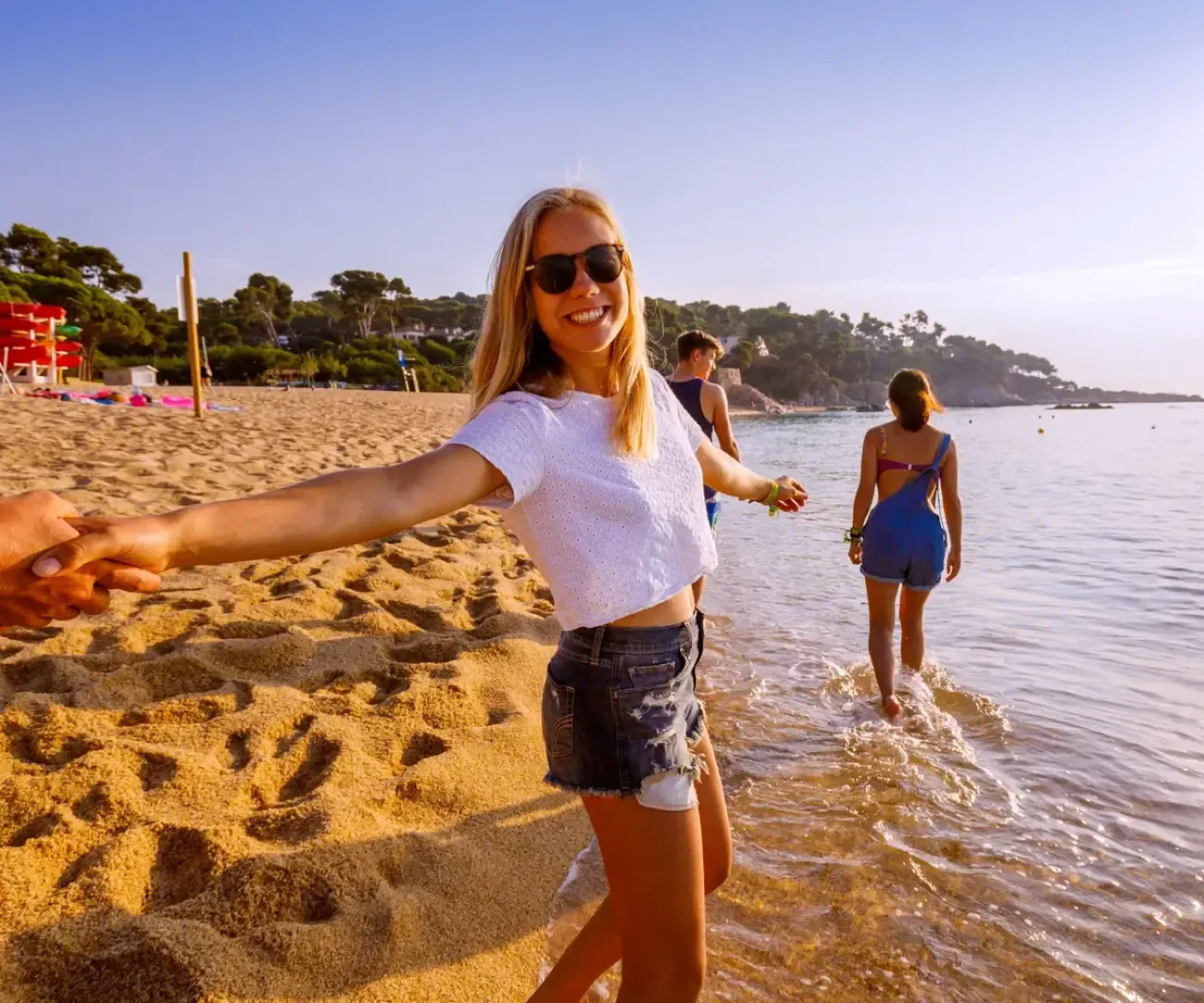 Eine lächelnde Frau mit Sonnenbrille und Shorts hält die Hand einer Person an einem Sandstrand, während zwei Personen im flachen Wasser spazieren gehen und im Hintergrund Bäume unter einem klaren Himmel zu sehen sind.