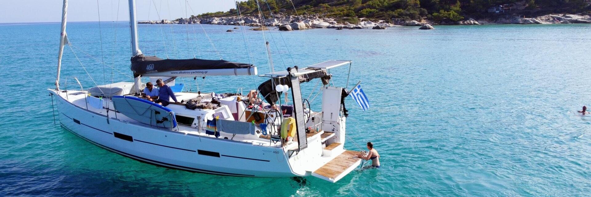 Ein weißes Segelboot mit Menschen an Bord ankert in klarem türkisfarbenem Wasser in der Nähe einer felsigen, von Bäumen gesäumten Küste. Eine griechische Flagge hängt am Boot, und eine Person schwimmt in der Nähe. Die Szene ist hell und sonnig.