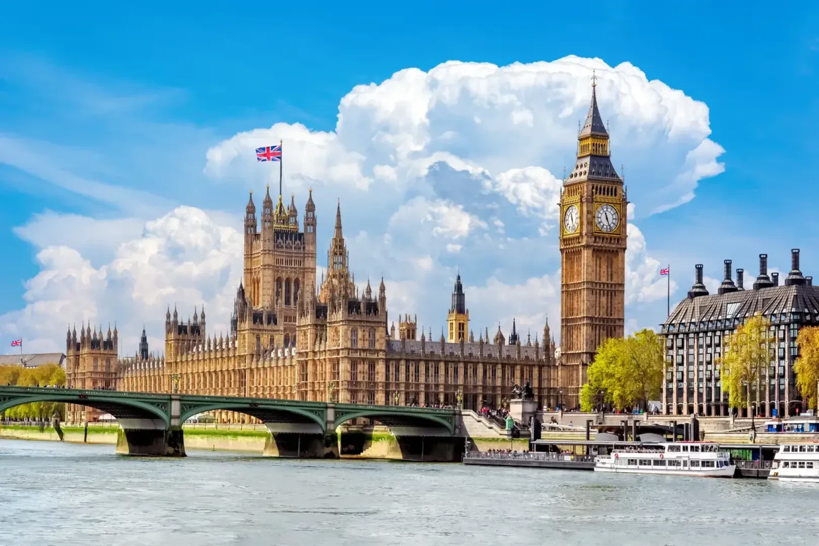 Die Houses of Parliament und der Uhrenturm Big Ben in London, Vereinigtes Königreich, mit wehender Union Jack-Flagge. Die Themse und die Westminster Bridge befinden sich im Vordergrund unter einem strahlend blauen Himmel mit großen Wolken.