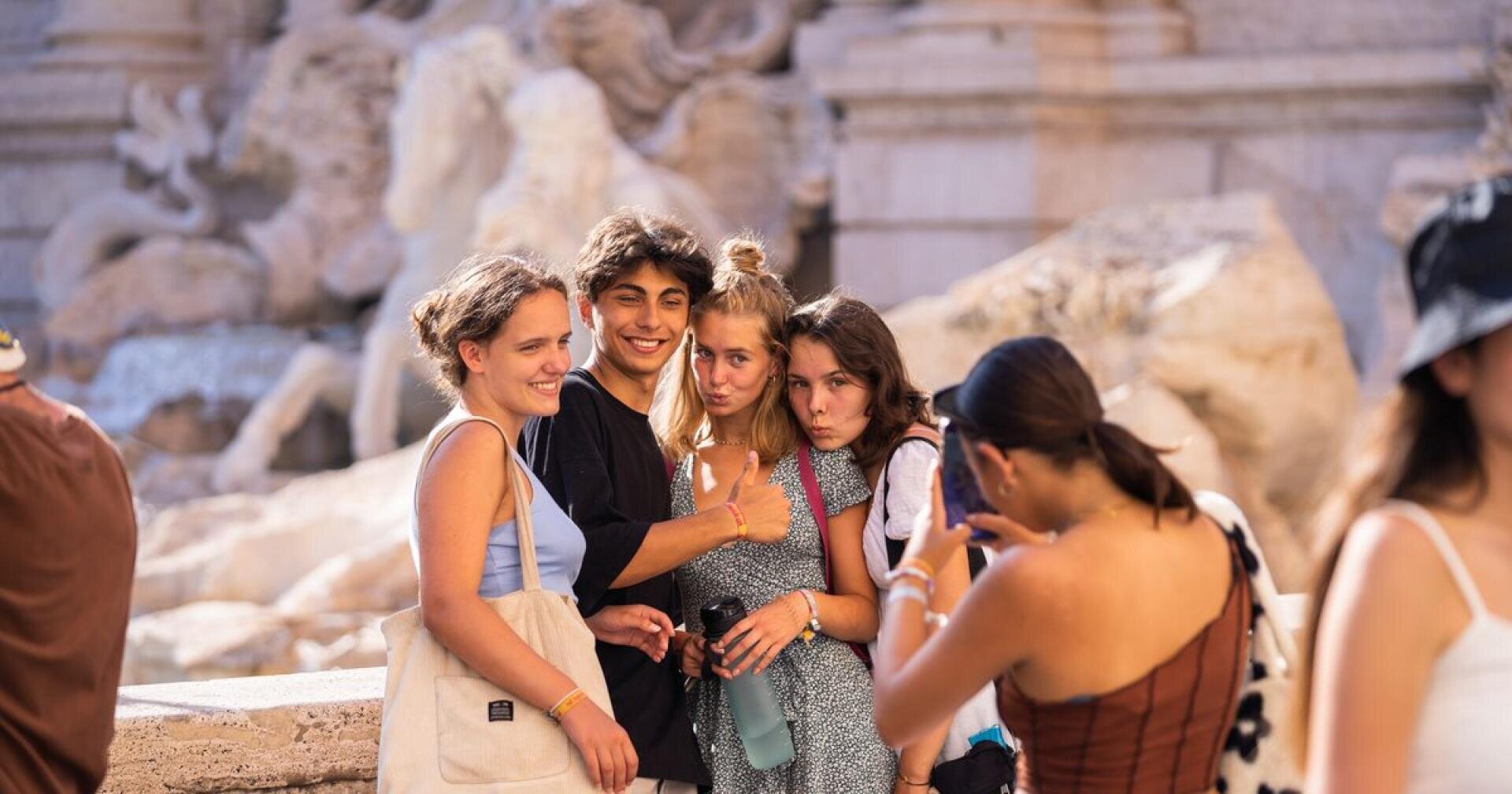 Eine Gruppe von vier jungen Leuten posiert eng beieinander lächelnd für ein Foto, während eine andere Person im Vordergrund an einer historischen Stätte im Freien mit Statuen ein Foto von ihnen macht.