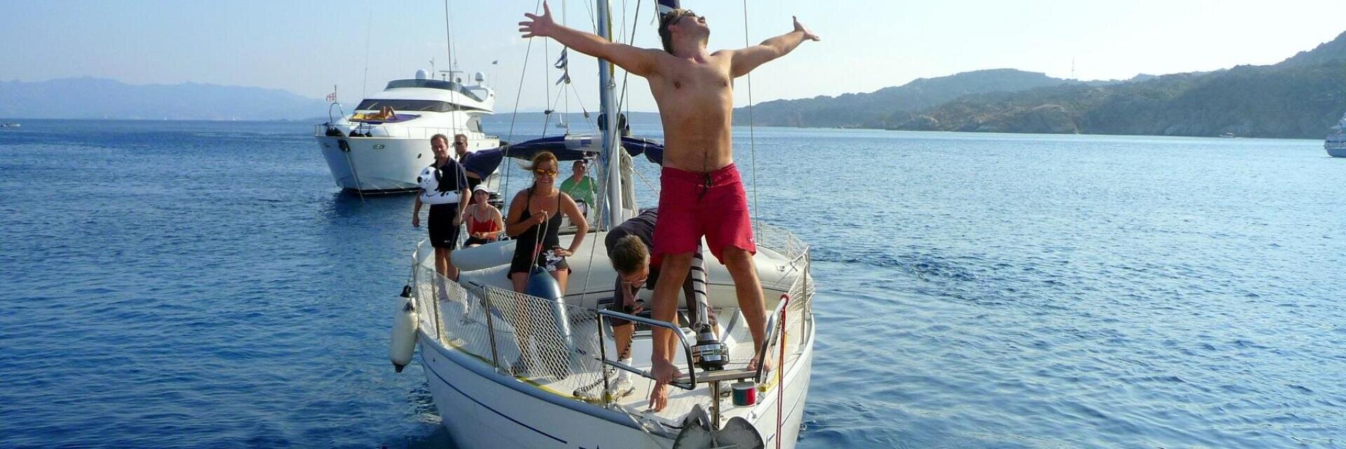 Eine Gruppe von Menschen entspannt sich auf einem Segelboot in ruhigem, blauem Wasser, mit einem hemdlosen Mann in roten Shorts, der mit ausgestreckten Armen am Bug steht. Im Hintergrund sind andere Boote und entfernte Hügel zu sehen.
