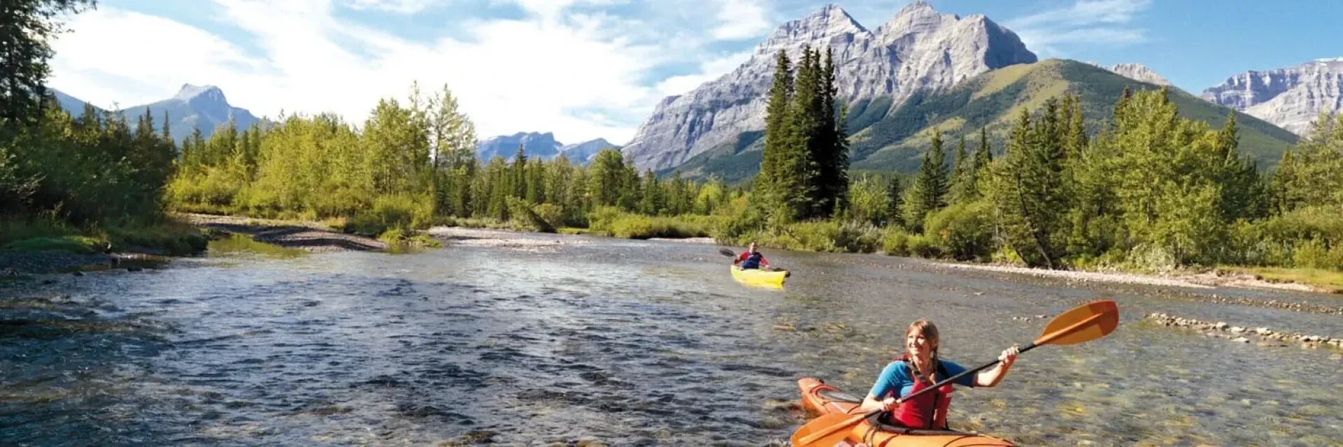Zwei Personen fahren mit dem Kajak auf einem klaren, flachen Fluss, umgeben von grünen Bäumen und Bergen unter einem blauen Himmel mit vereinzelten Wolken.