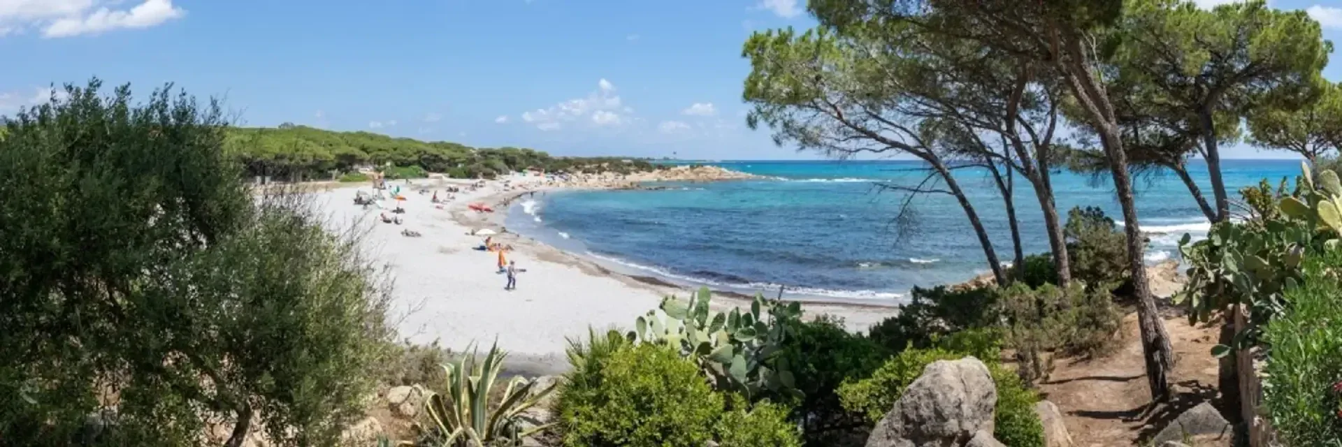 Ein Panoramablick auf einen Sandstrand mit Sonnenschirmen und Menschen, türkisfarbenen Wellen und üppig grünen Bäumen und Sträuchern entlang des Ufers unter einem teilweise bewölkten Himmel.