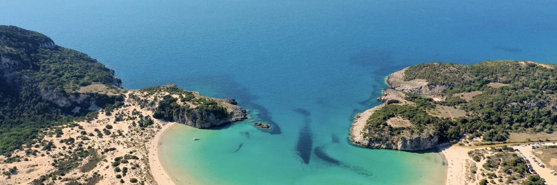 Luftaufnahme einer türkisfarbenen Bucht, umgeben von Sandstränden und zerklüfteten, grünen Hügeln, mit kristallklarem, blauem Meer, das sich bis zum Horizont erstreckt.