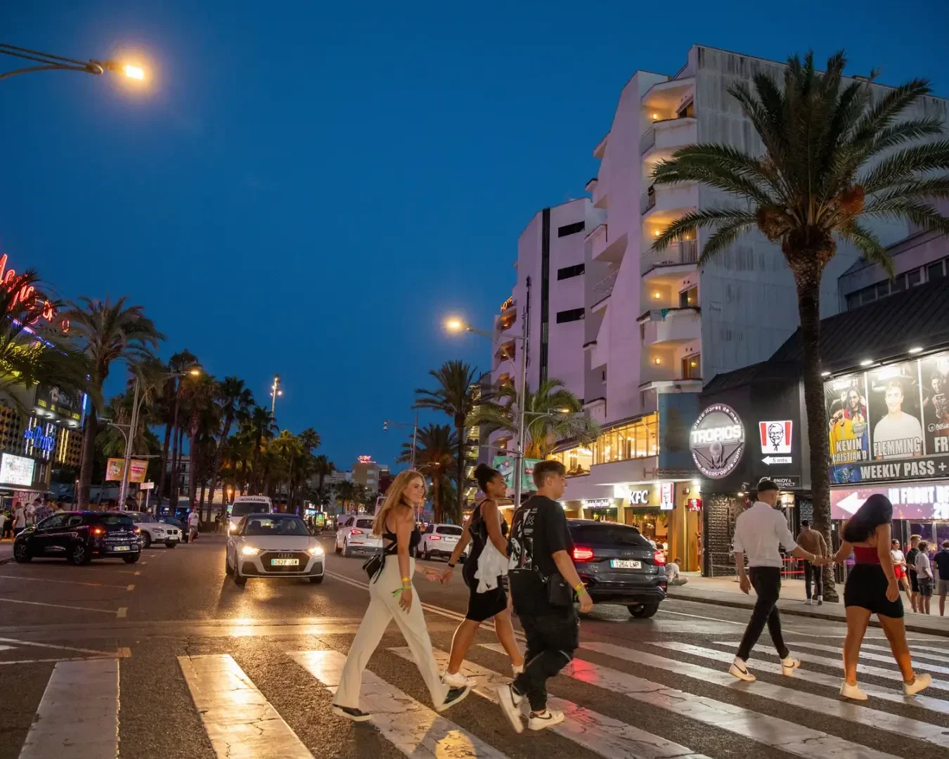 Vier junge Erwachsene überqueren in der Abenddämmerung eine belebte Straße in der Stadt, umgeben von Palmen, Autos und beleuchteten Gebäuden mit Leuchtreklamen und Werbetafeln. Der Himmel ist tiefblau, und die Szene ist lebendig und pulsierend.