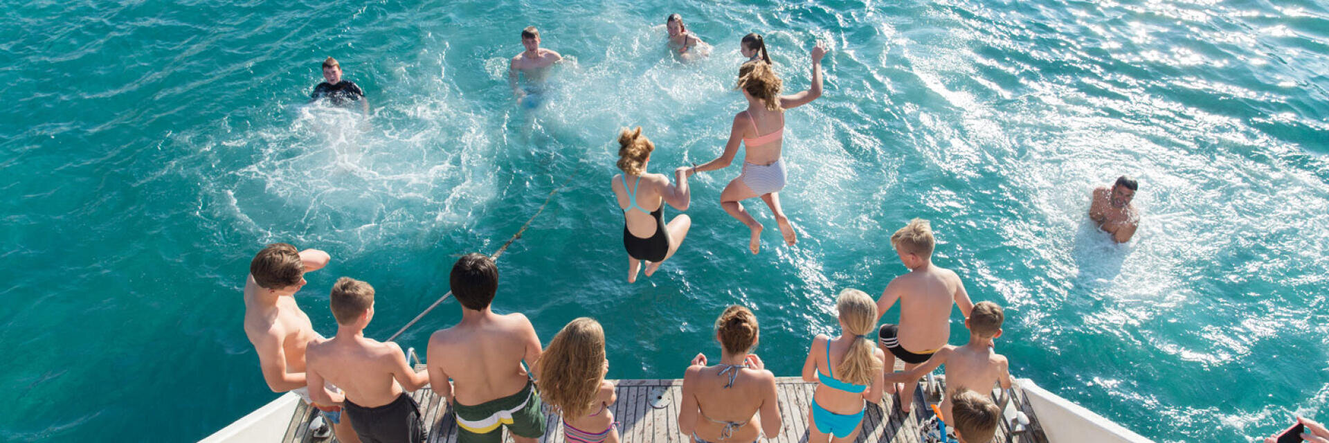 Eine Gruppe von Menschen, meist Kinder und Jugendliche in Badeanzügen, springt von einem Boot in das klare blaue Wasser, während andere in der Nähe schwimmen; die Szene ist lebhaft und fröhlich im hellen Sonnenlicht.