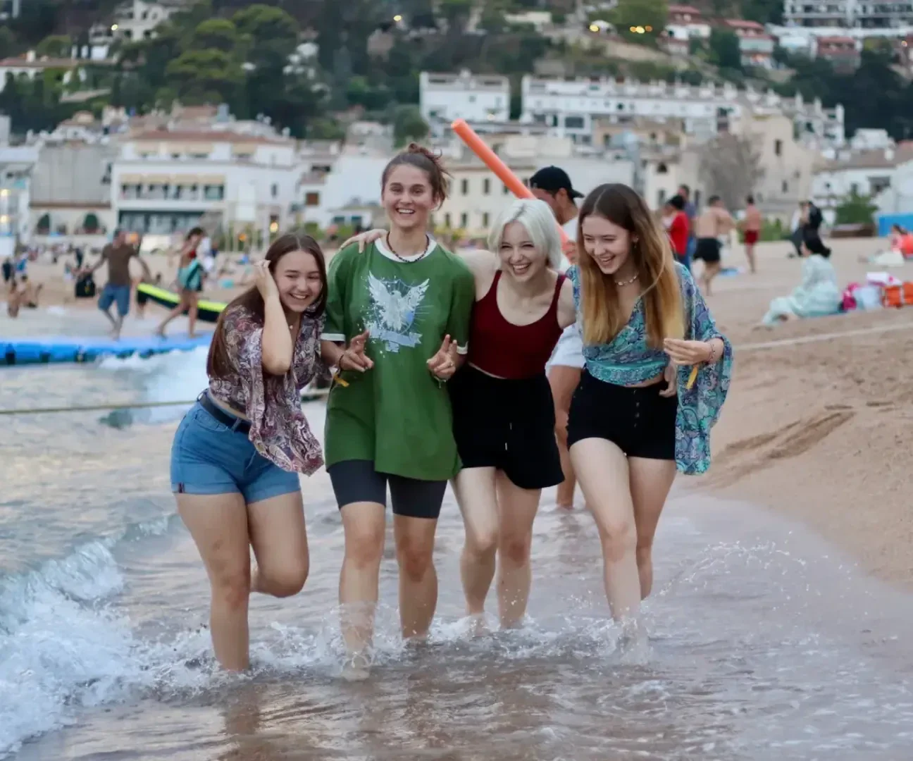 Vier junge Frauen spazieren lächelnd und lachend durch das flache Wasser am Strand, während im Hintergrund eine belebte Küste, Gebäude und grüne Hügel zu sehen sind.