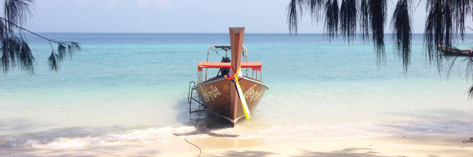 Ein Holzboot mit einem orangefarbenen Band liegt an einem Sandstrand mit sanften Wellen vor Anker. Das klare türkisfarbene Meer erstreckt sich bis zum Horizont, und Kiefernzweige umrahmen den oberen Bildrand.