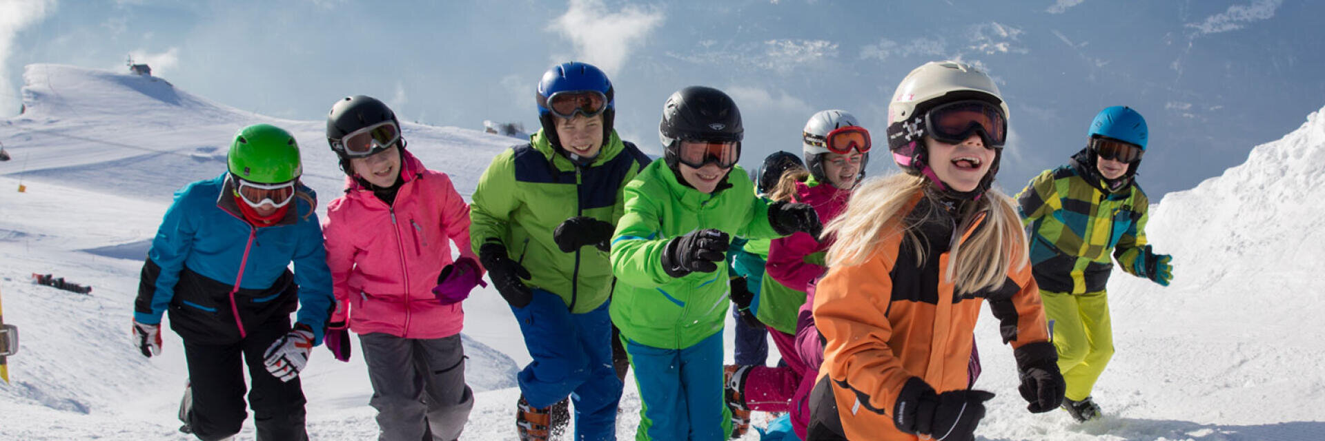 Eine Gruppe von Kindern in bunten Winterklamotten und mit Helmen rennt an einem sonnigen Tag fröhlich einen verschneiten Berghang hinauf, mit schneebedeckten Gipfeln und einem blauen Himmel im Hintergrund.